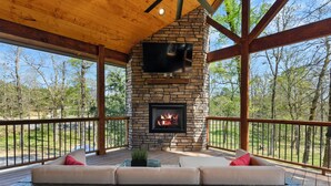 A covered outdoor area with a stone fireplace, a sofa, and a television mounted on the wall. - The Hickory Nut | 2 Bed | 2.5 Bath | Sleeps 4 (Broken Bow)
