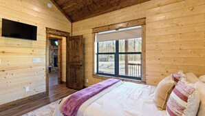 A wooden cabin bedroom with a bed, pillows, a TV mounted on the wall, and a window. - The Hickory Nut | 2 Bed | 2.5 Bath | Sleeps 4 (Broken Bow)