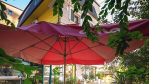 An outdoor patio with round tables and chairs, covered by a red umbrella, surrounded by potted plants and a building in the background. - Hotel Aria (Tirana)