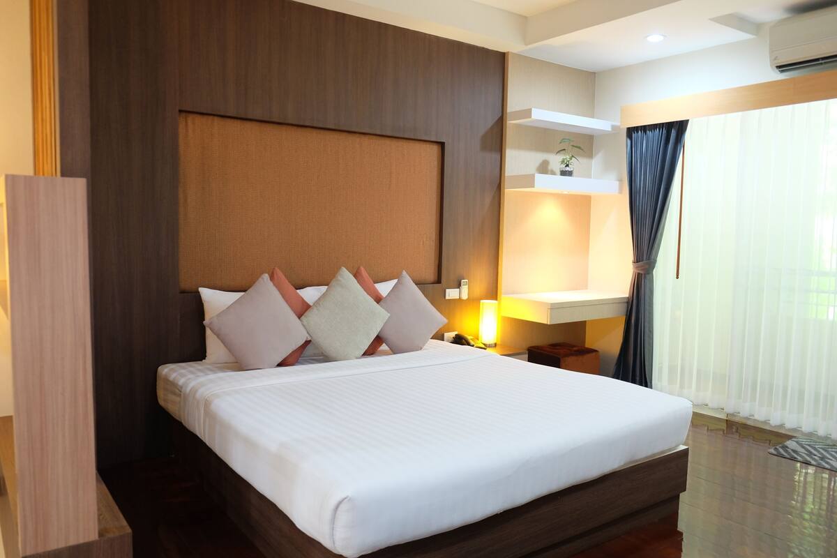 Una cama bien hecha con cabecera de madera, una mesita de noche con lámpara y una ventana con cortinas.