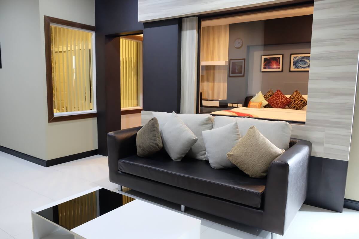 Una sala de estar moderna con un sofá oscuro, cojines blancos y una mesa de centro de vidrio. Se ve un área de cocina al fondo con una mesa de comedor y sillas.