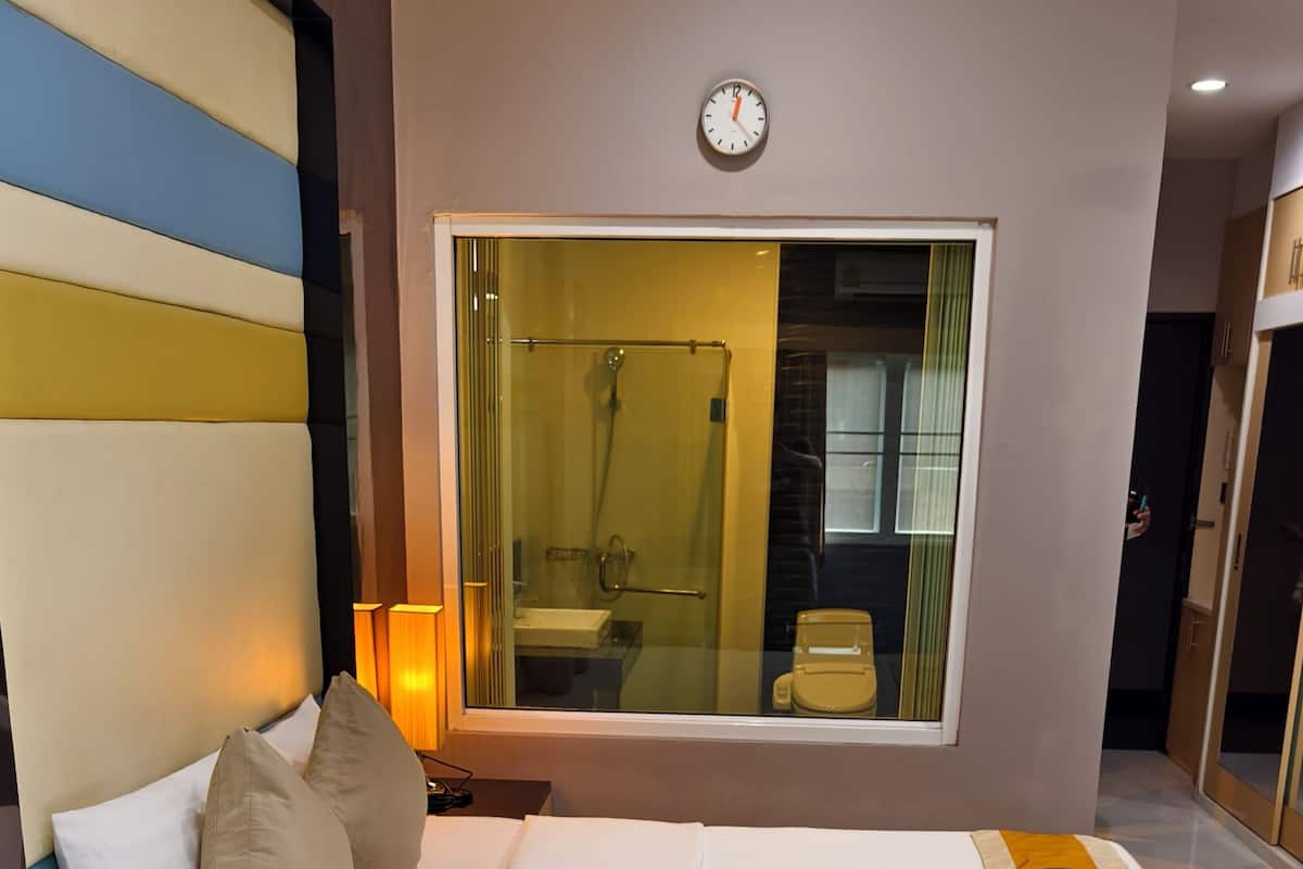 Habitación de hotel con una cama grande, una mesita de noche con un teléfono, una ventana con vistas al baño y un reloj en la pared.