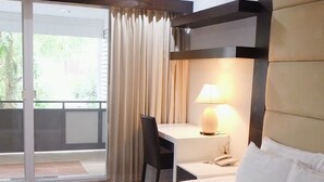 A hotel room with a bed, a desk with a lamp, a chair, and a view of greenery outside. - Eastpana 304(R1) Executive Residence (Si Maha Phot)