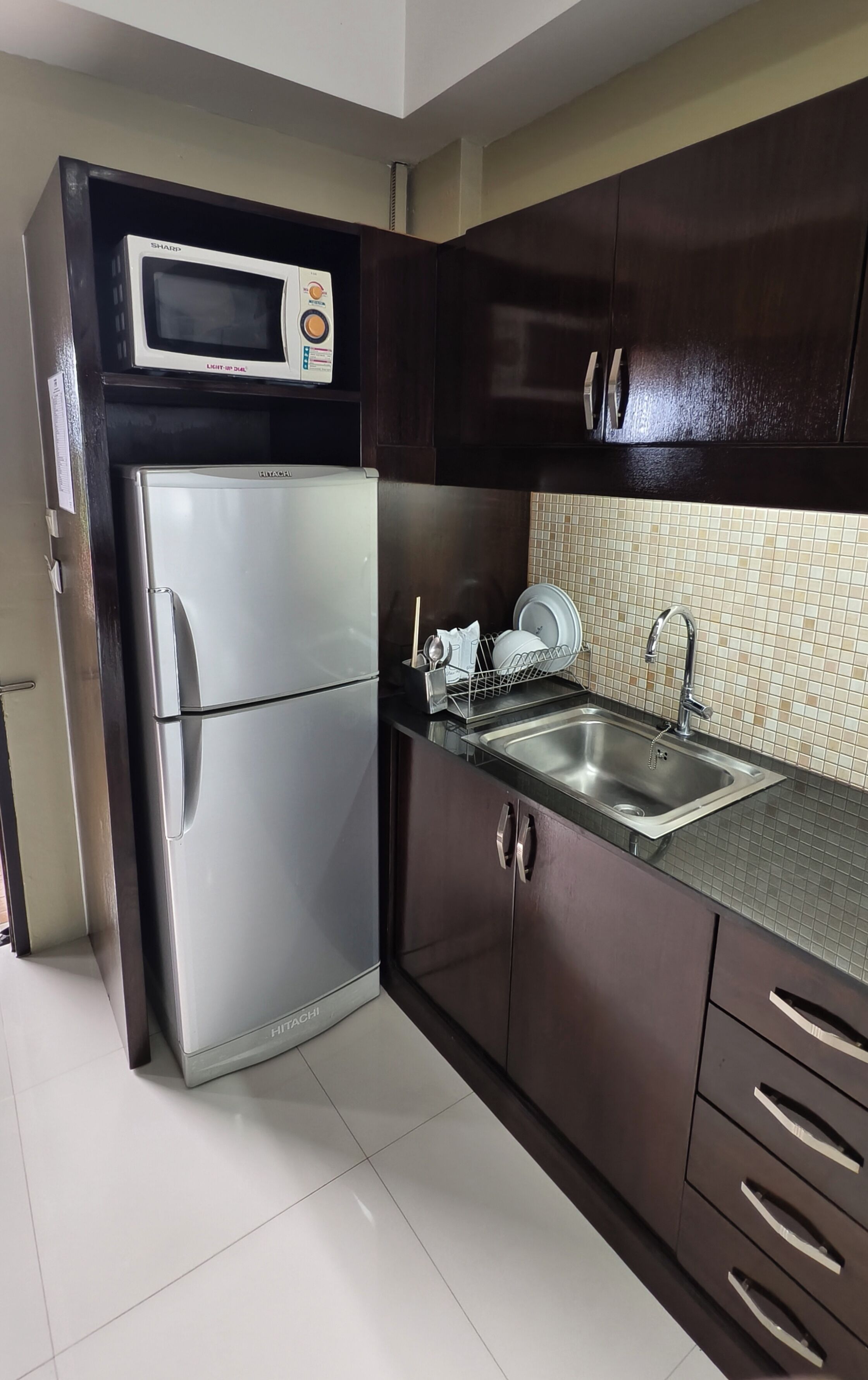 Cocina compacta con microondas integrado, refrigerador de acero inoxidable, pileta con grifo moderno y alacenas de madera oscura.