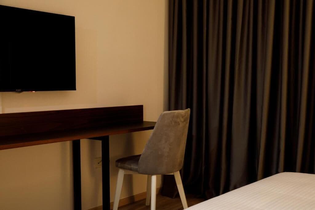 A hotel room with a bed, a desk, a chair, a flat-screen TV mounted on the wall, and a window with dark curtains.