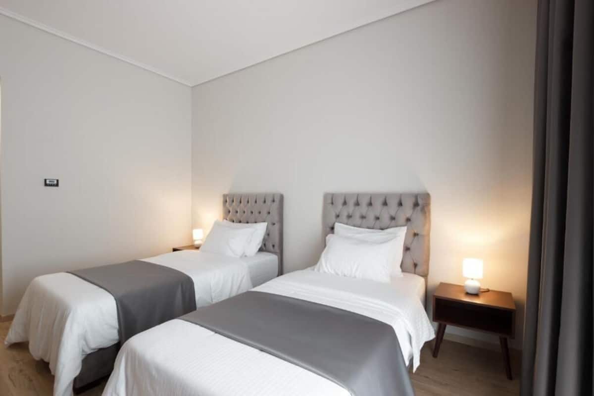 A hotel room with two beds, each with a grey bedspread and white pillows, a tufted headboard, and a bedside table with a lamp.