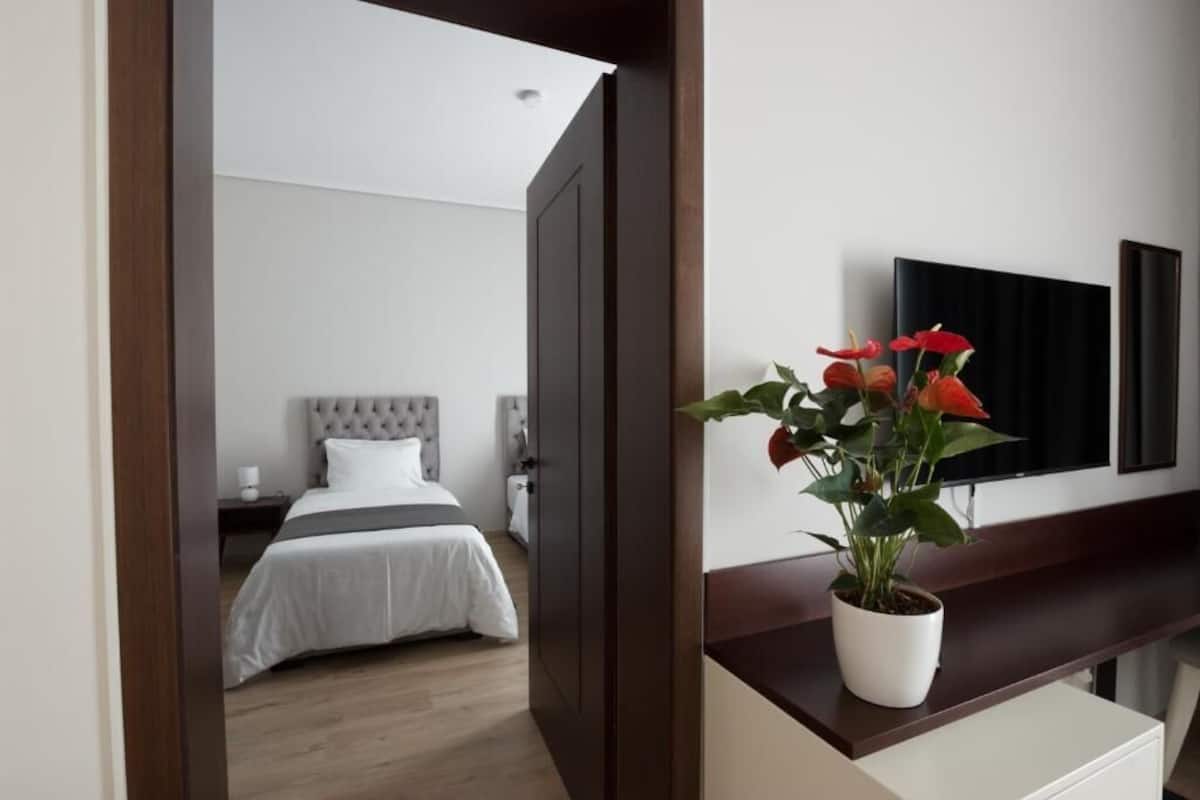 A modern hotel room with a bed, a bedside table, a TV mounted on the wall, and a potted plant on a wooden console.