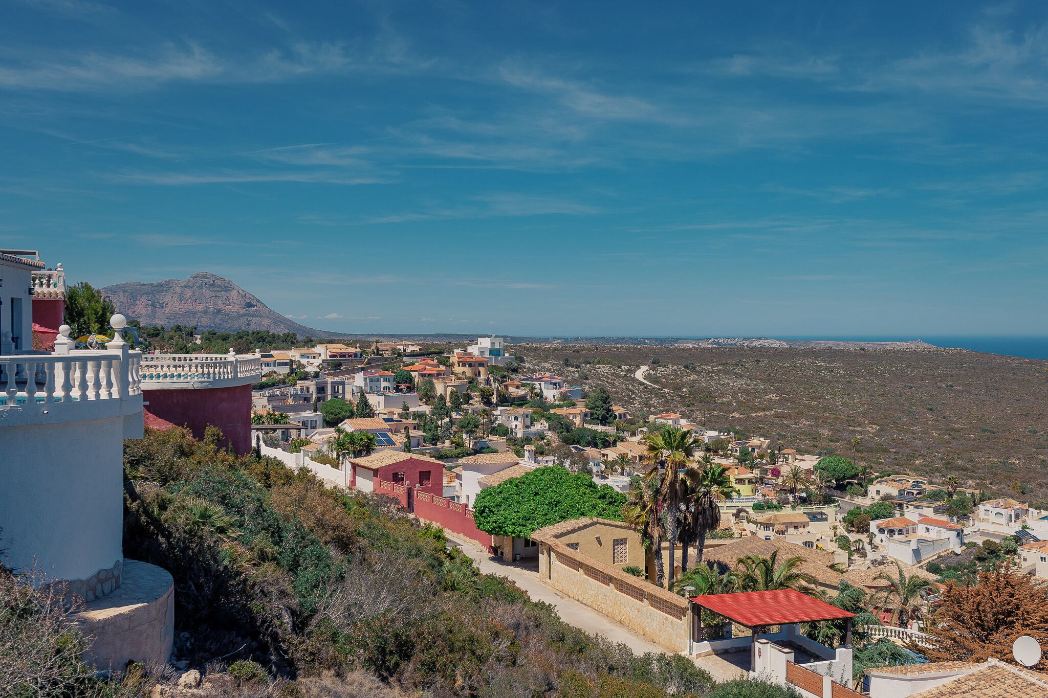 A coastal town with a prominent mountain in the background, terraced houses, and a clear blue sky.