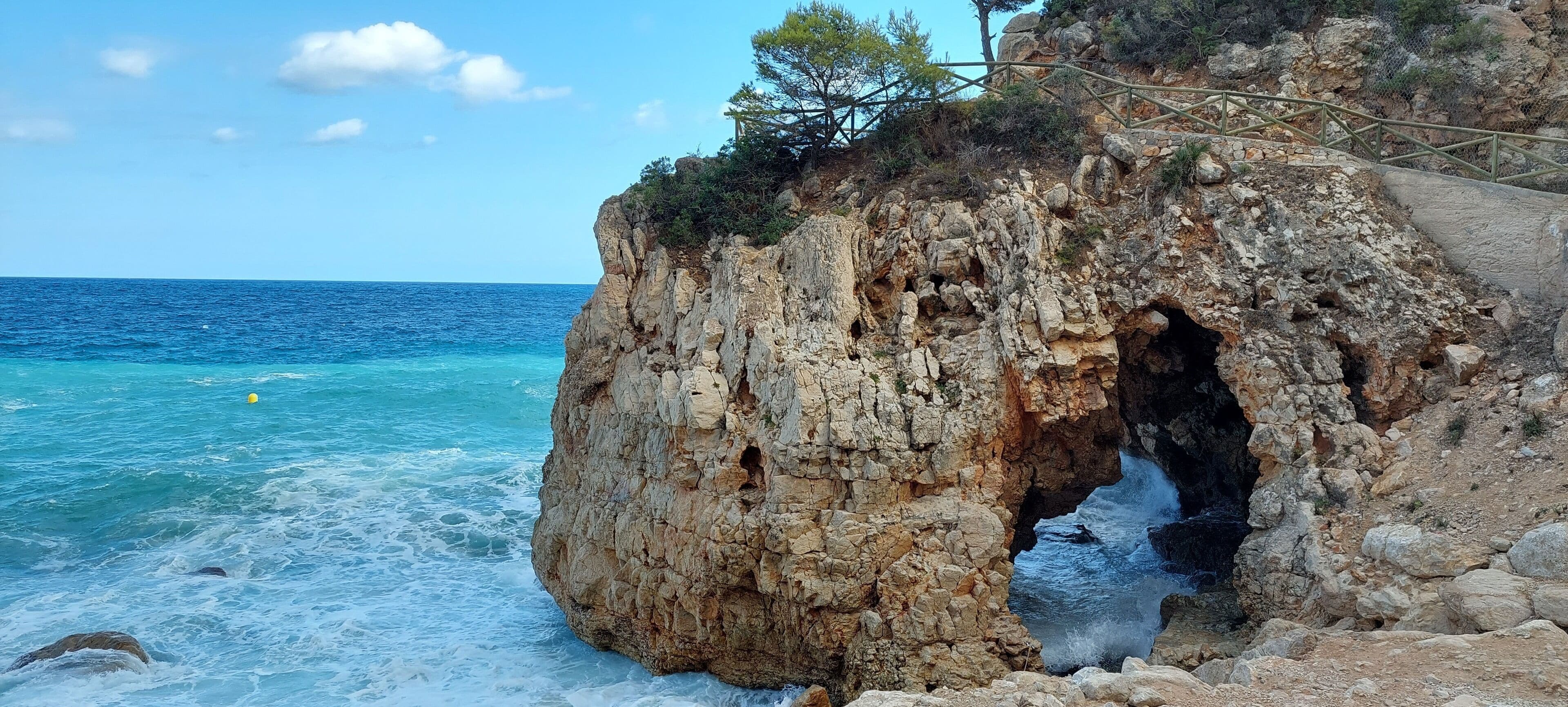 Un paysage côtier rocheux avec l’entrée d’une grotte, une mer d’un bleu limpide et un escalier en bois menant vers le haut.