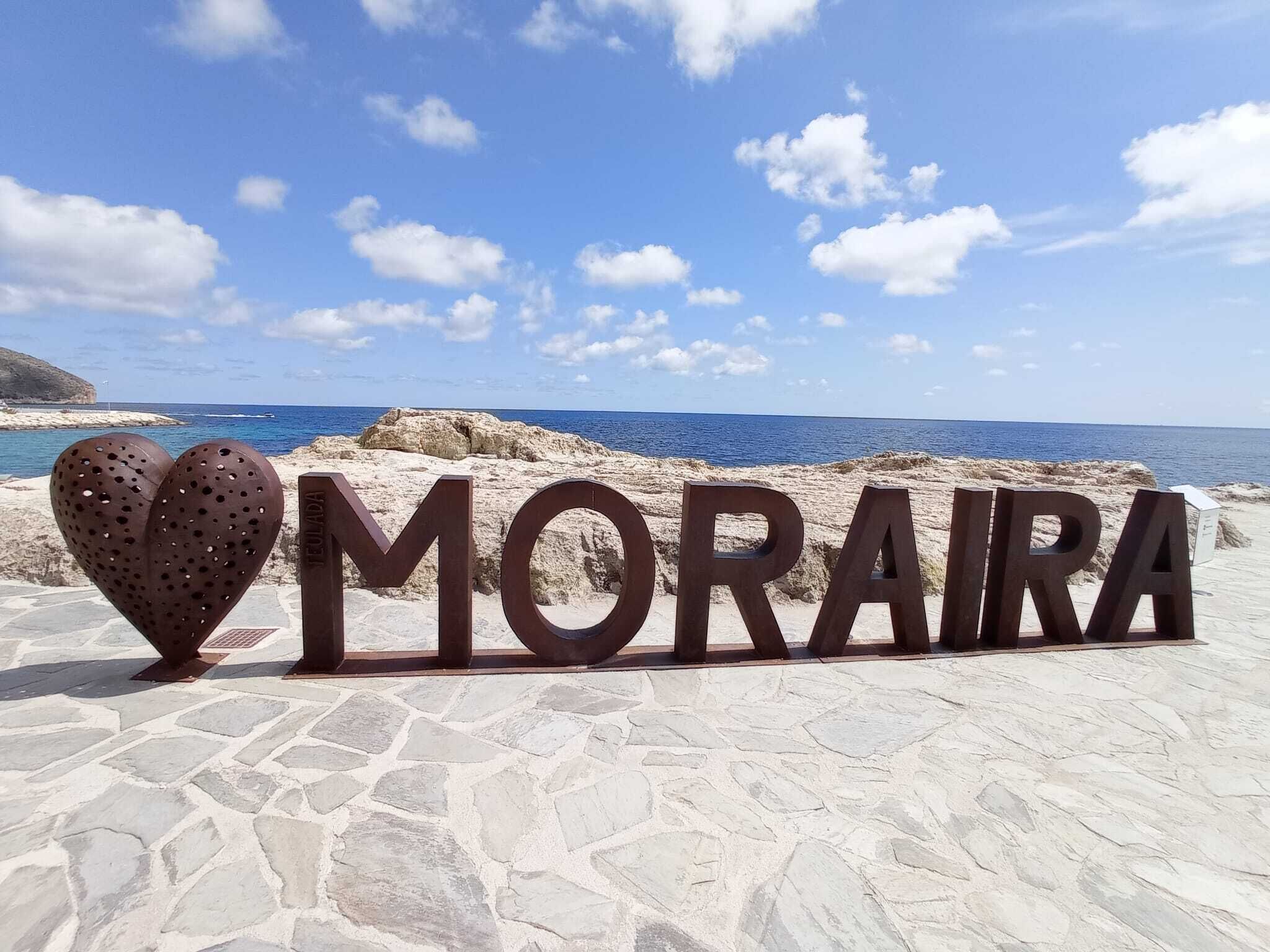 A sign with the word "MORARA" and a heart-shaped object in front of a coastal landscape.
