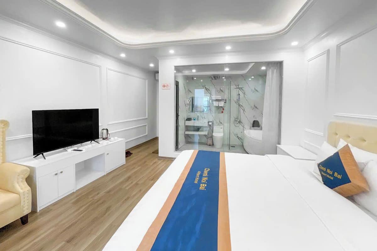 A modern hotel room with a large bed, a TV, a desk, and a bathroom visible through a glass door.