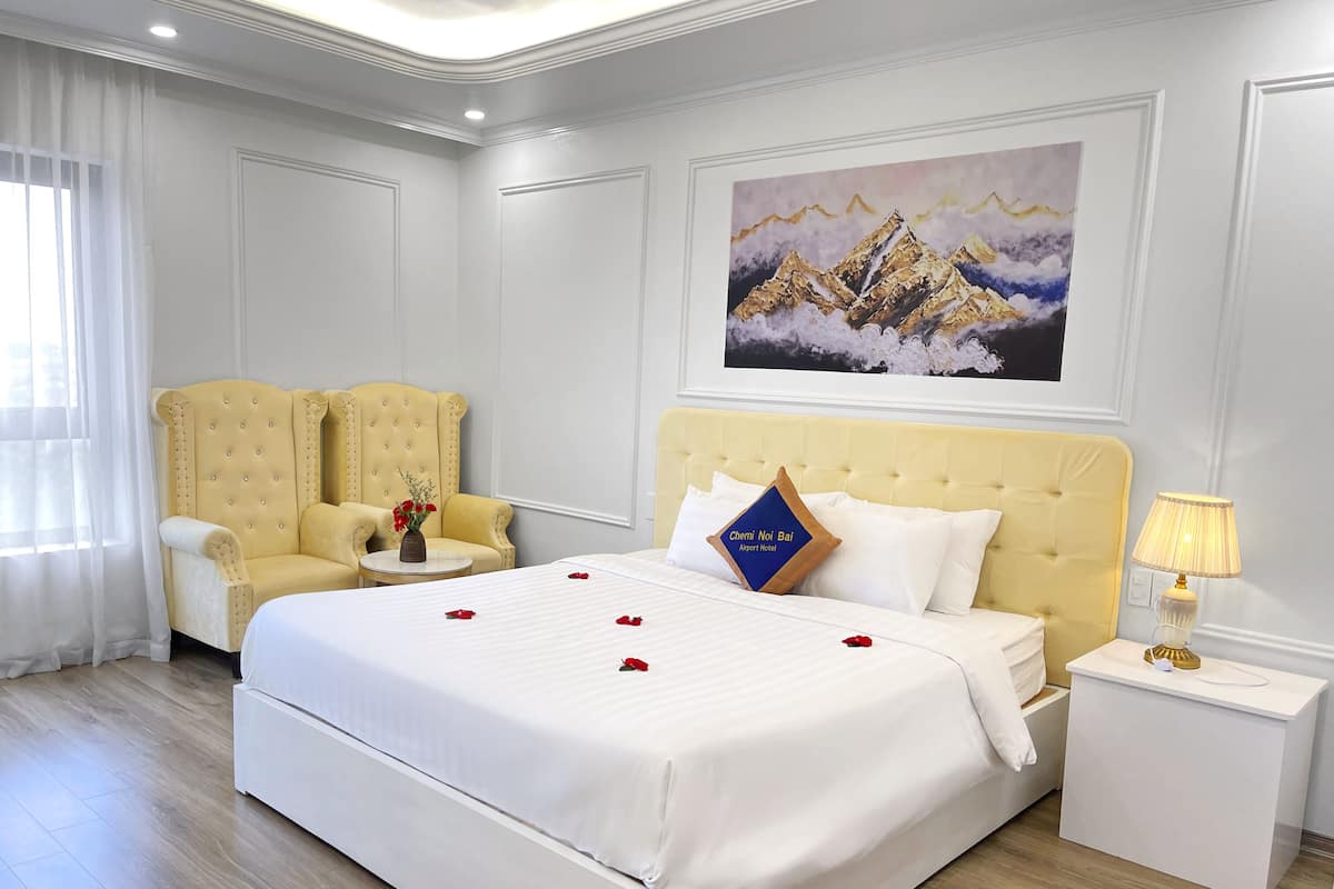 A hotel room with a large bed, two armchairs, a small table with a vase, a painting on the wall, and a nightstand with a lamp.