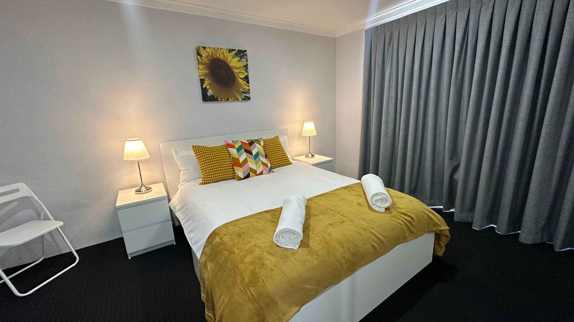 Habitació d'hotel amb un llit, dues taules de nit, una cadira, un quadre de girasol i cortines grises.