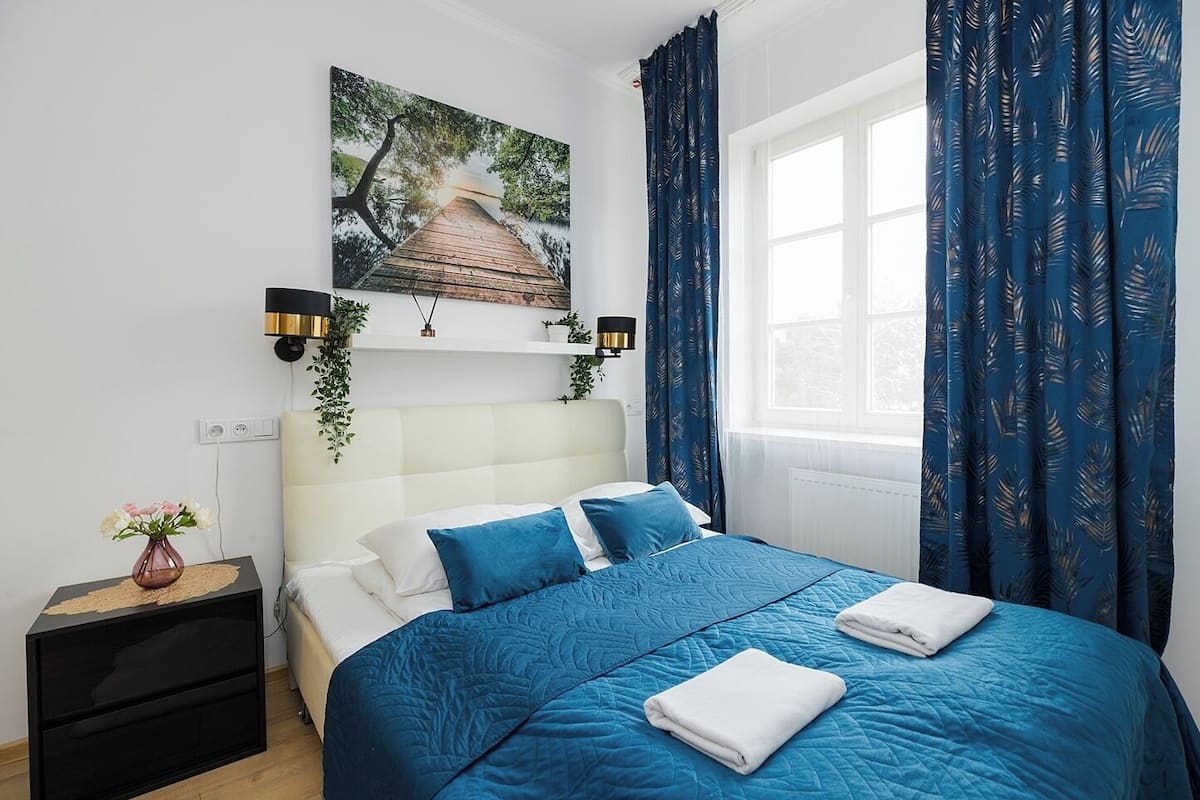 Quarto com cama, mesa de cabeceira, prateleira na parede com plantas e janela com cortinas.