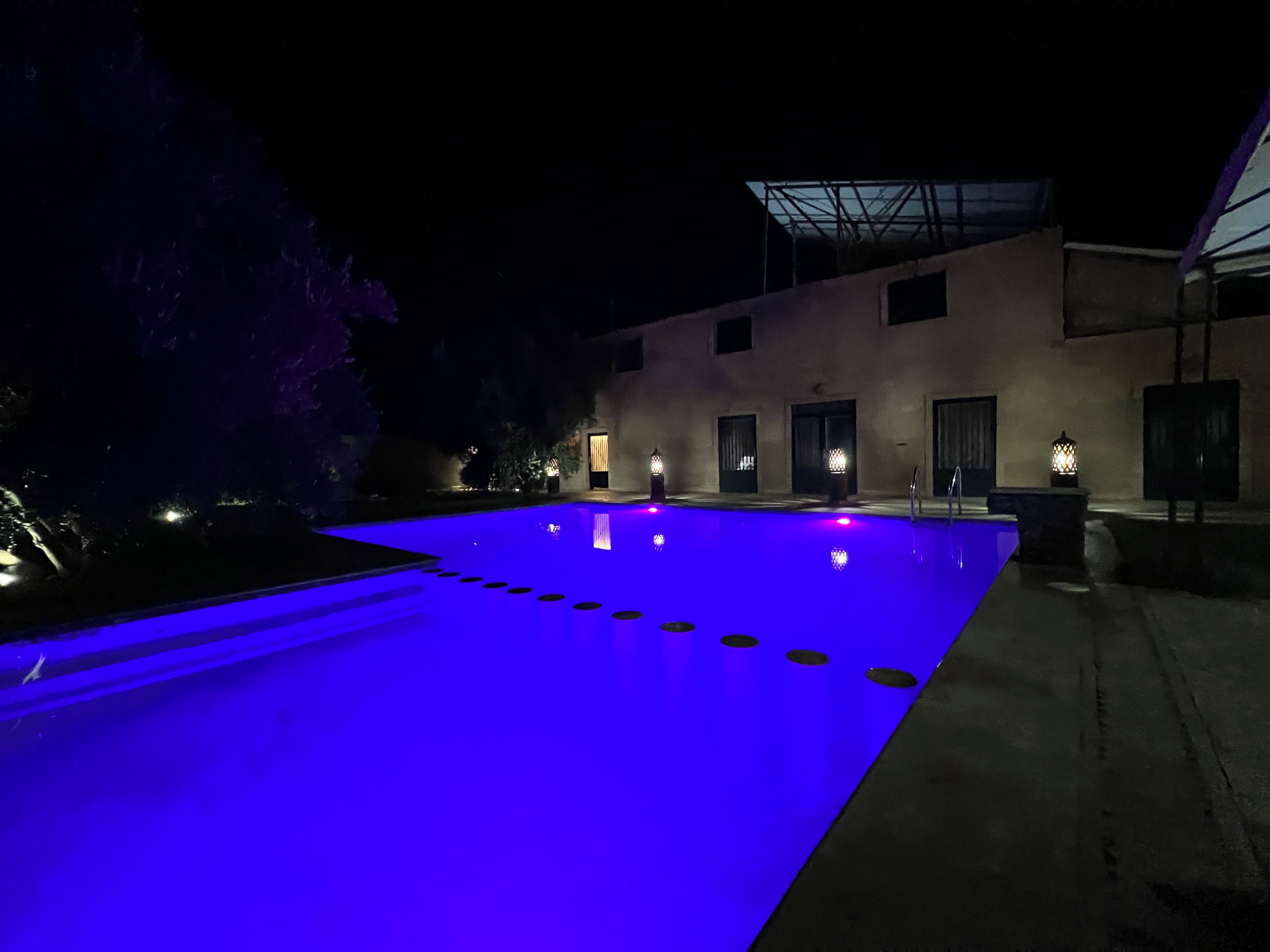 Une piscine avec une terrasse illuminée de lumière bleue, un bâtiment doté de nombreuses portes et un éclairage extérieur.