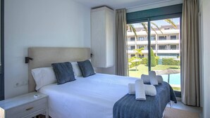A hotel room with a large bed, a nightstand, a window with a view of a pool and buildings, and a ceiling light. - Samos Apartment (Badia de Xàbia)