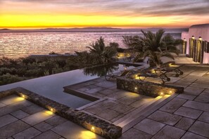 A rooftop infinity pool with stone borders and ambient lighting, overlooking a sunset over the sea.