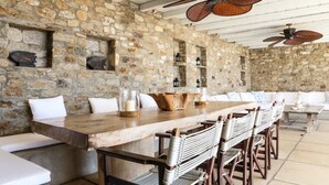 A stone-walled dining area with a long wooden table, wicker chairs, and ceiling fans. - Casa di Mare: Luxury and Breathtaking Views in Mykonos (Mykonos)