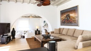 A modern living room with a large sofa, a coffee table, and a television. There is a painting on the wall and a ceiling fan. - Casa di Mare: Luxury and Breathtaking Views in Mykonos (Mykonos)