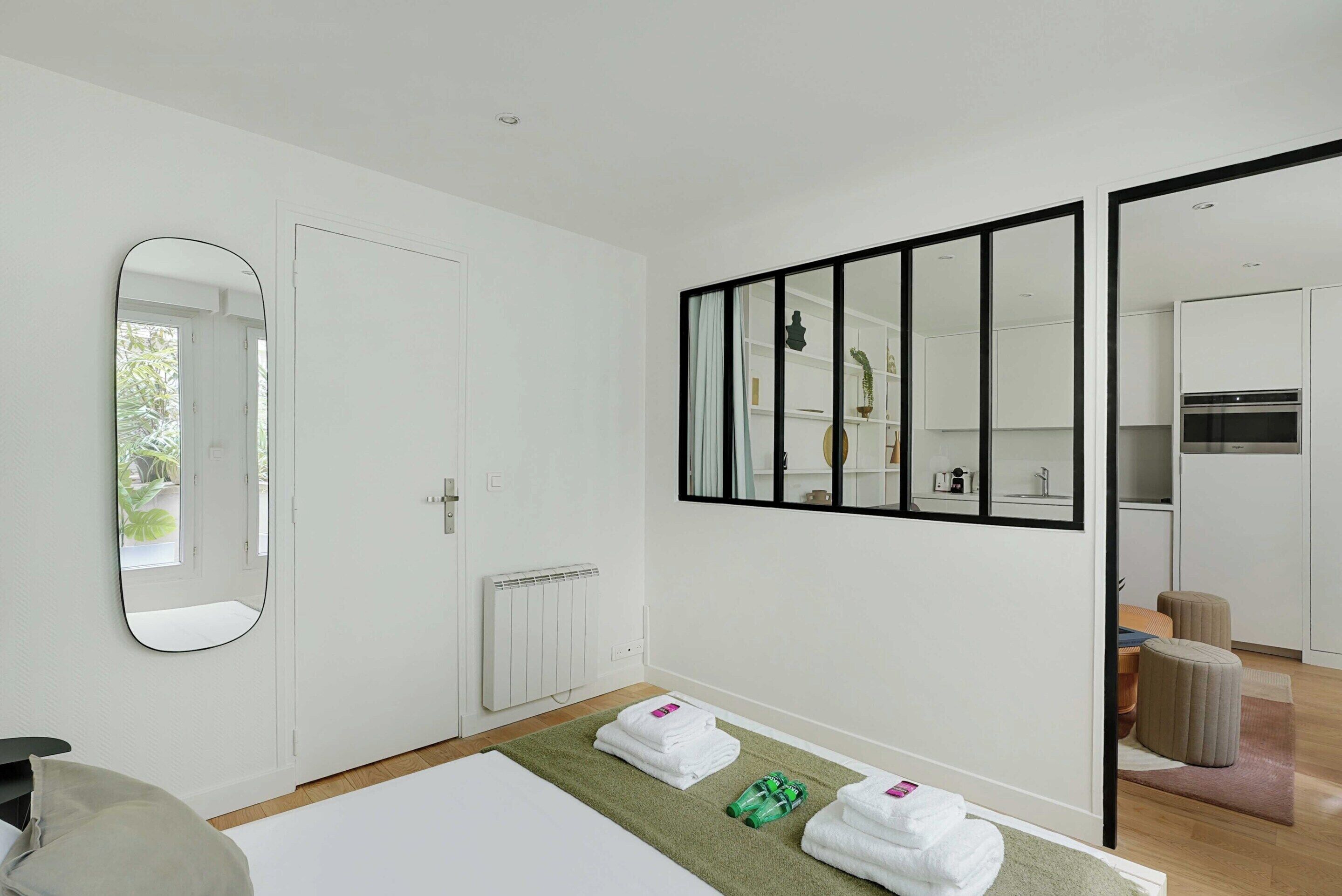 Habitación de hotel moderna con un espejo grande, una cama bien hecha, una alfombra verde y vistas a la cocina a través de una puerta corredera.