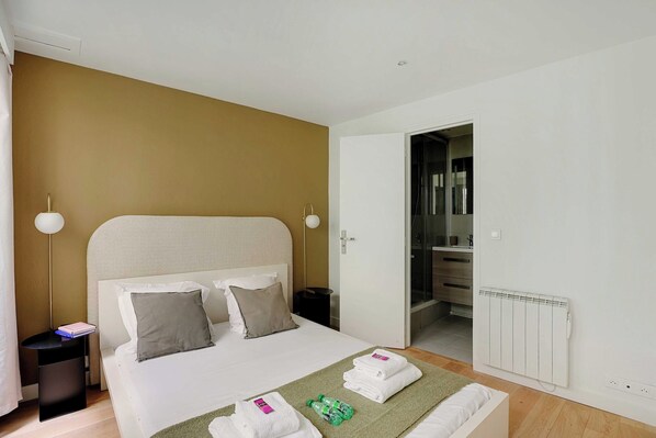 A hotel room with a bed, bedside tables, a radiator, and a bathroom visible through an open door.