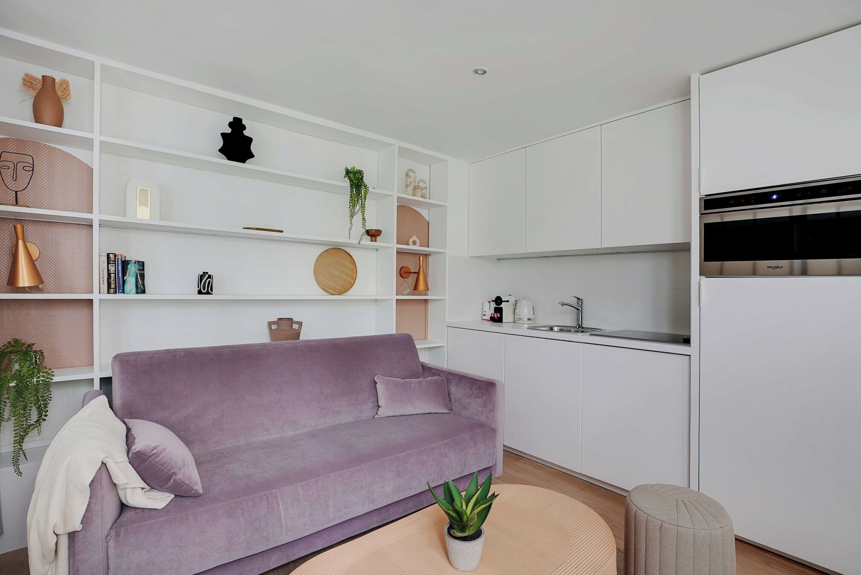 A modern living room with a purple sofa, white shelving with decorative items, a kitchen area with white cabinets and appliances, and a round wooden table.
