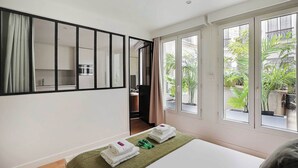 A hotel room with a bed, a green area rug, a large window with a view of greenery, and a mirror with a black frame.