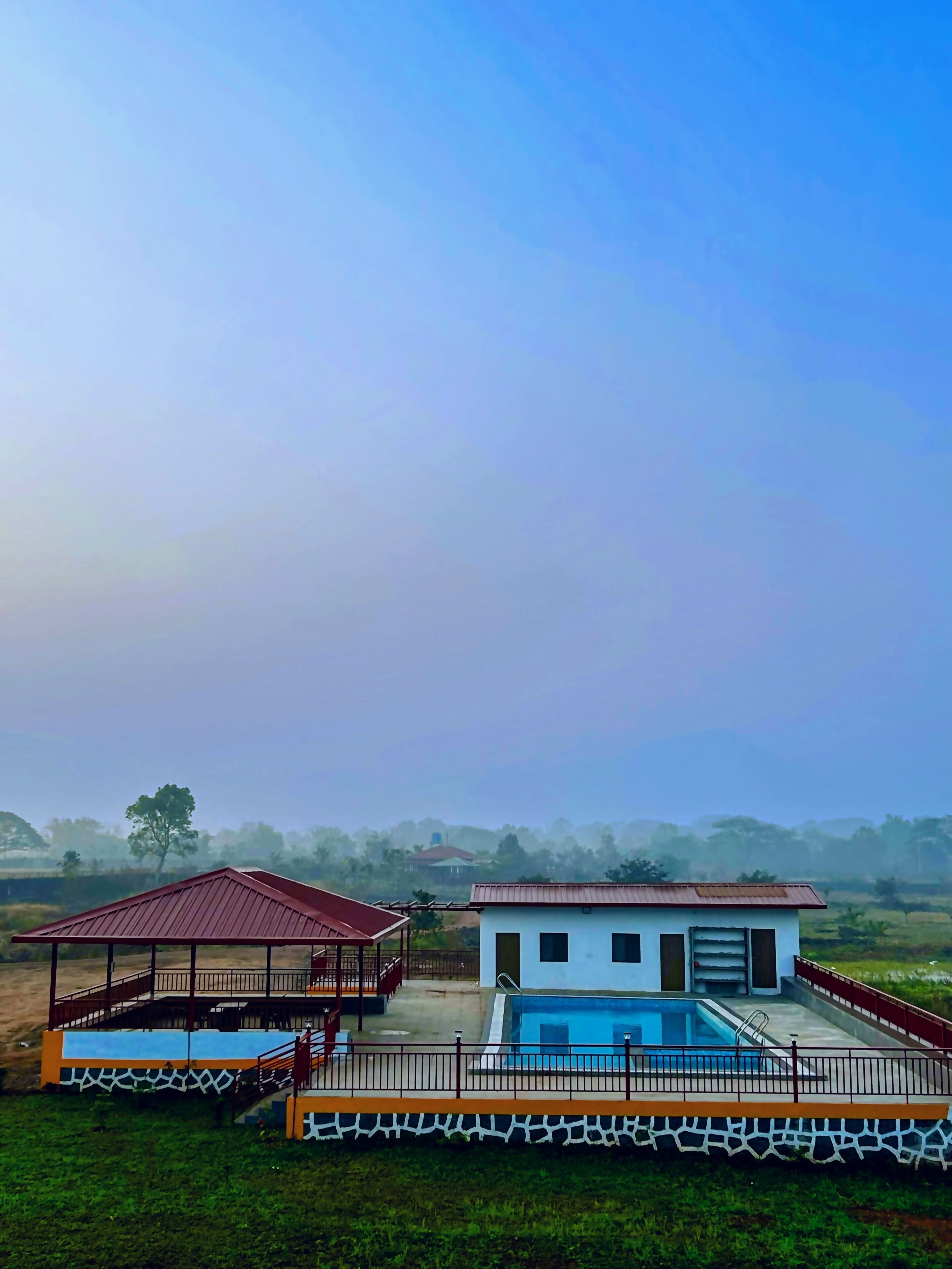 Área de piscina com um pavilhão coberto e um pequeno edifício, cercada por vegetação e um céu claro.