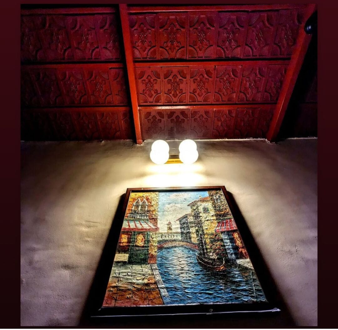 A framed painting of a bridge over water, placed on a table in a room with red patterned walls and two spherical light fixtures above.