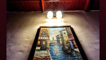 A framed painting of a bridge over water, placed on a table in a room with red patterned walls and two spherical light fixtures above.
