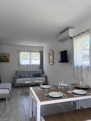 A modern living room with a sofa, a dining table set for two, and a TV mounted on the wall. - Maxima Sunset KONTIKI 4 pers (Sainte-Maxime)