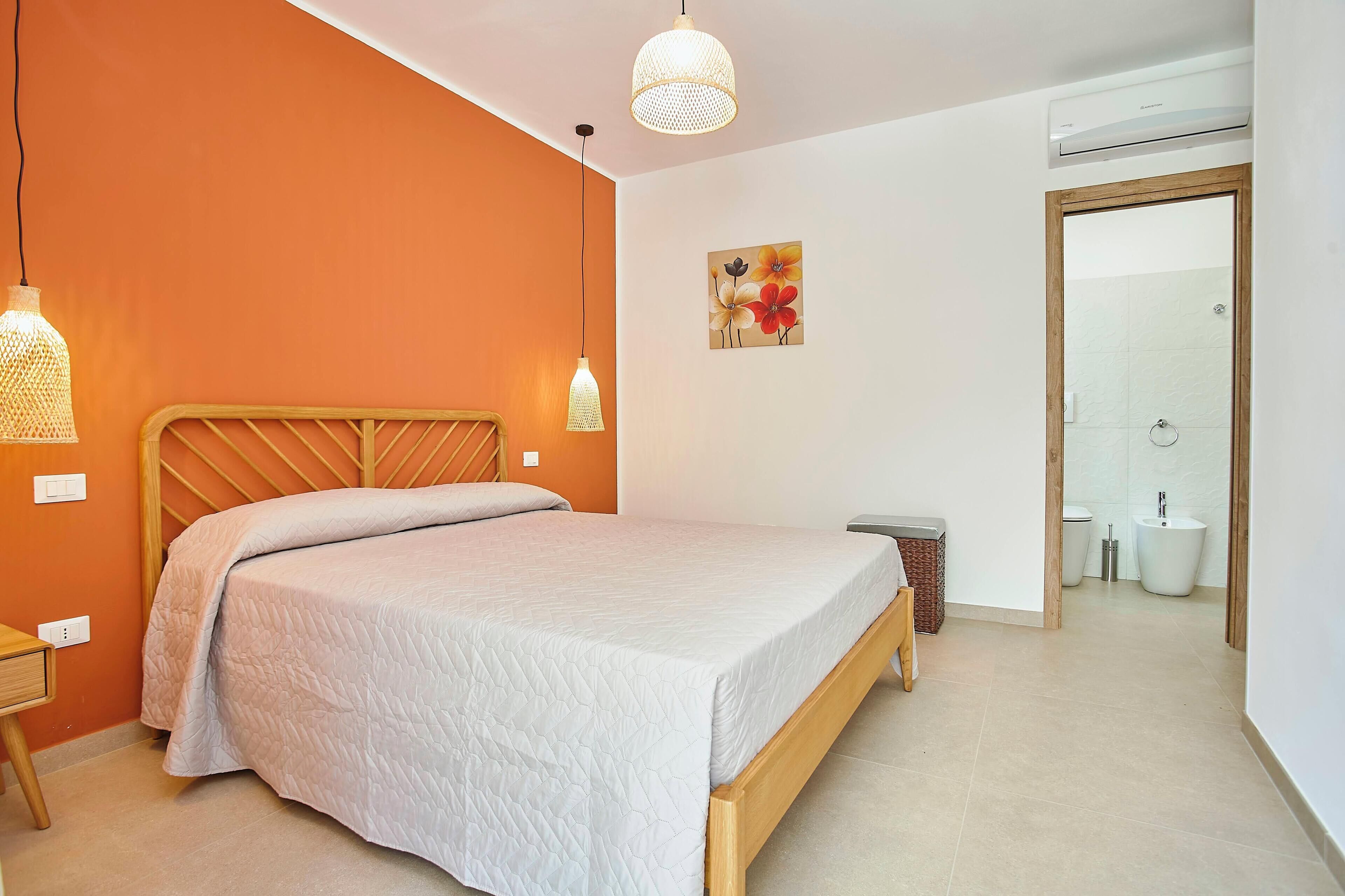 A bedroom with a bed, bedside table, hanging light, and a bathroom visible through an open door.