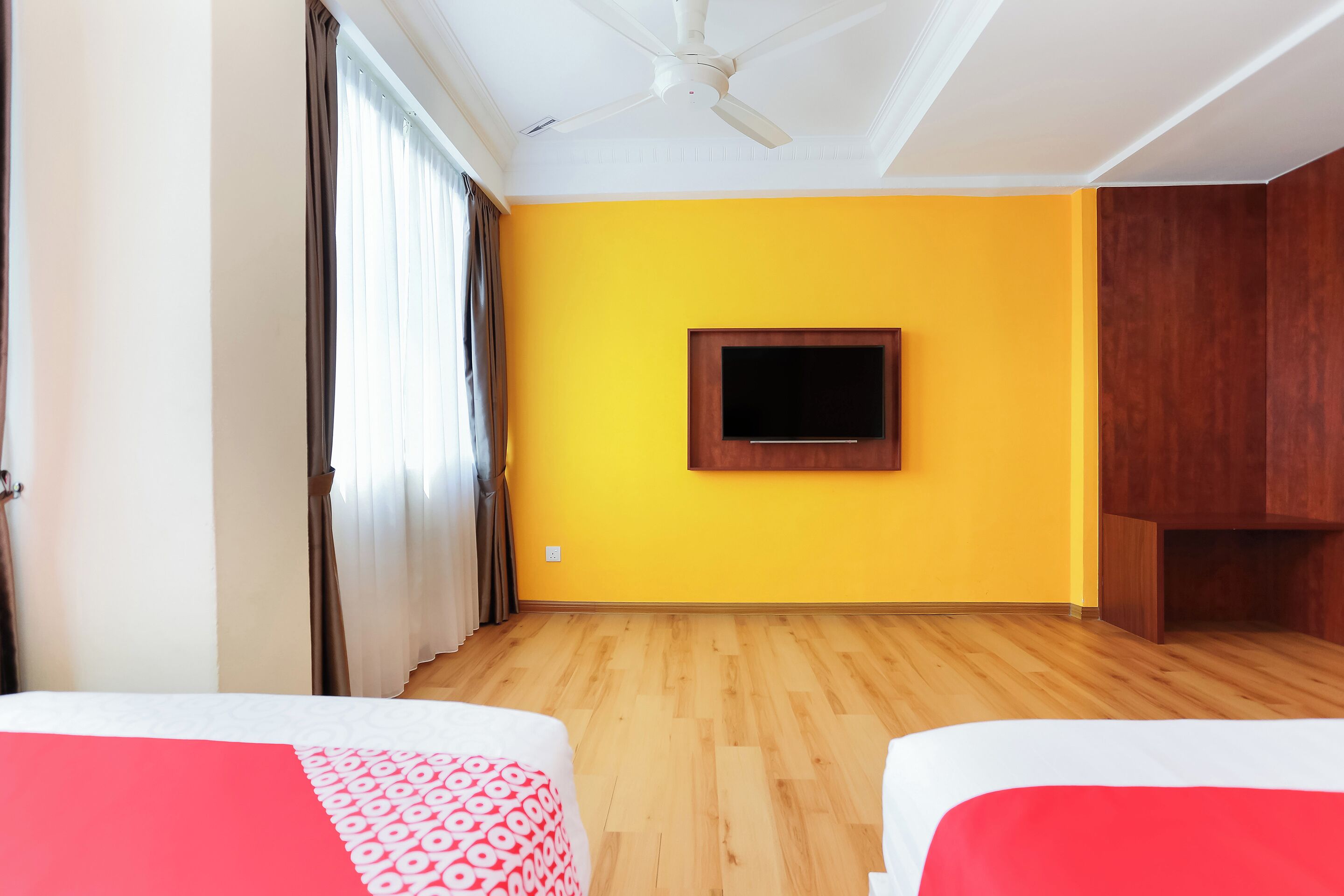 A room with two beds, a ceiling fan, a mounted TV, and wooden flooring.