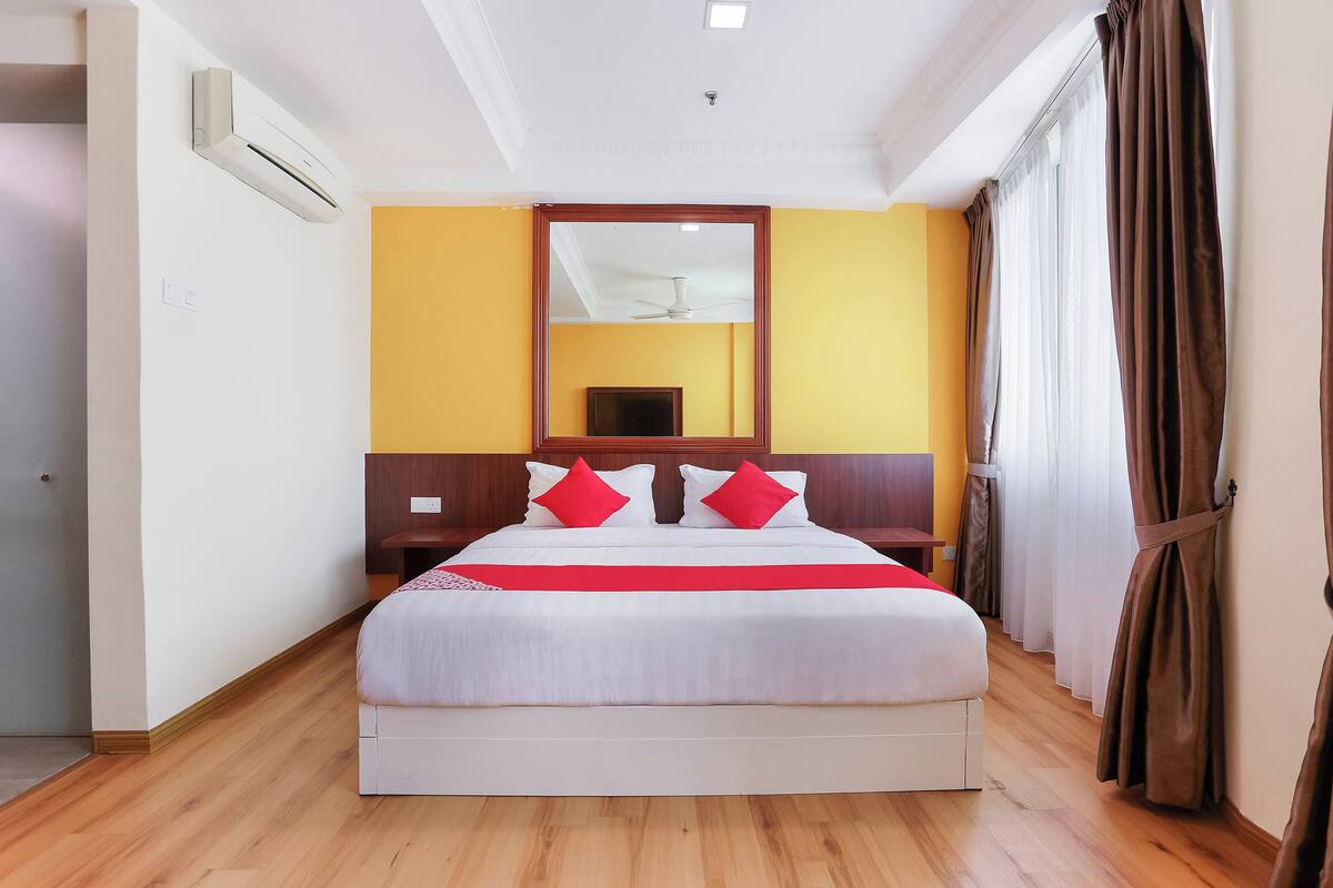 A hotel room with a large bed, a wooden headboard, a mirror above the bed, a ceiling fan, and a window with curtains.