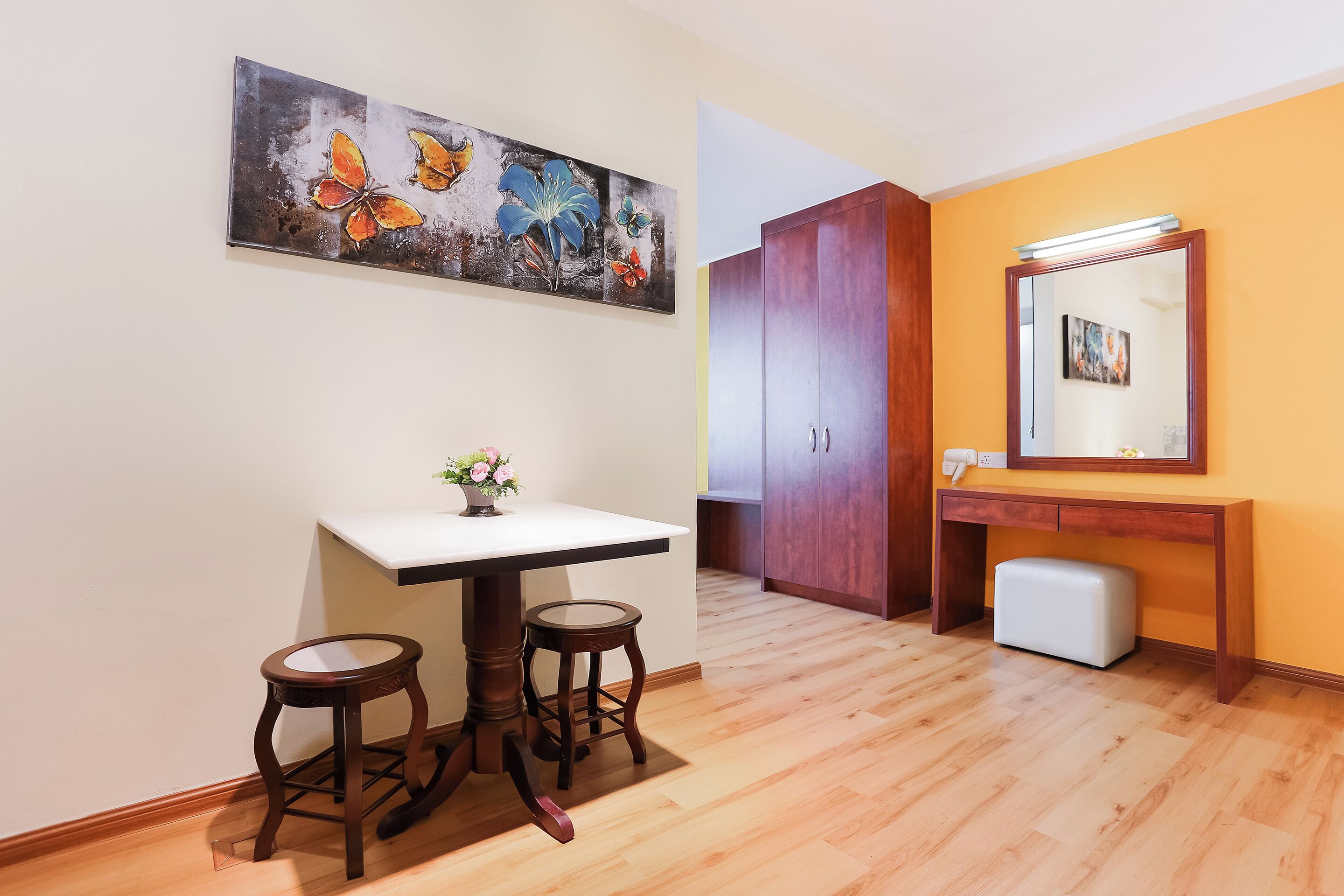 Una habitación con piso de madera, un escritorio blanco con dos taburetes, un armario de madera y un espejo con luz superior.