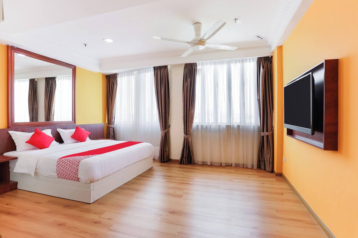 A hotel room with a large bed, a ceiling fan, a flat-screen TV, and a large window with curtains.
