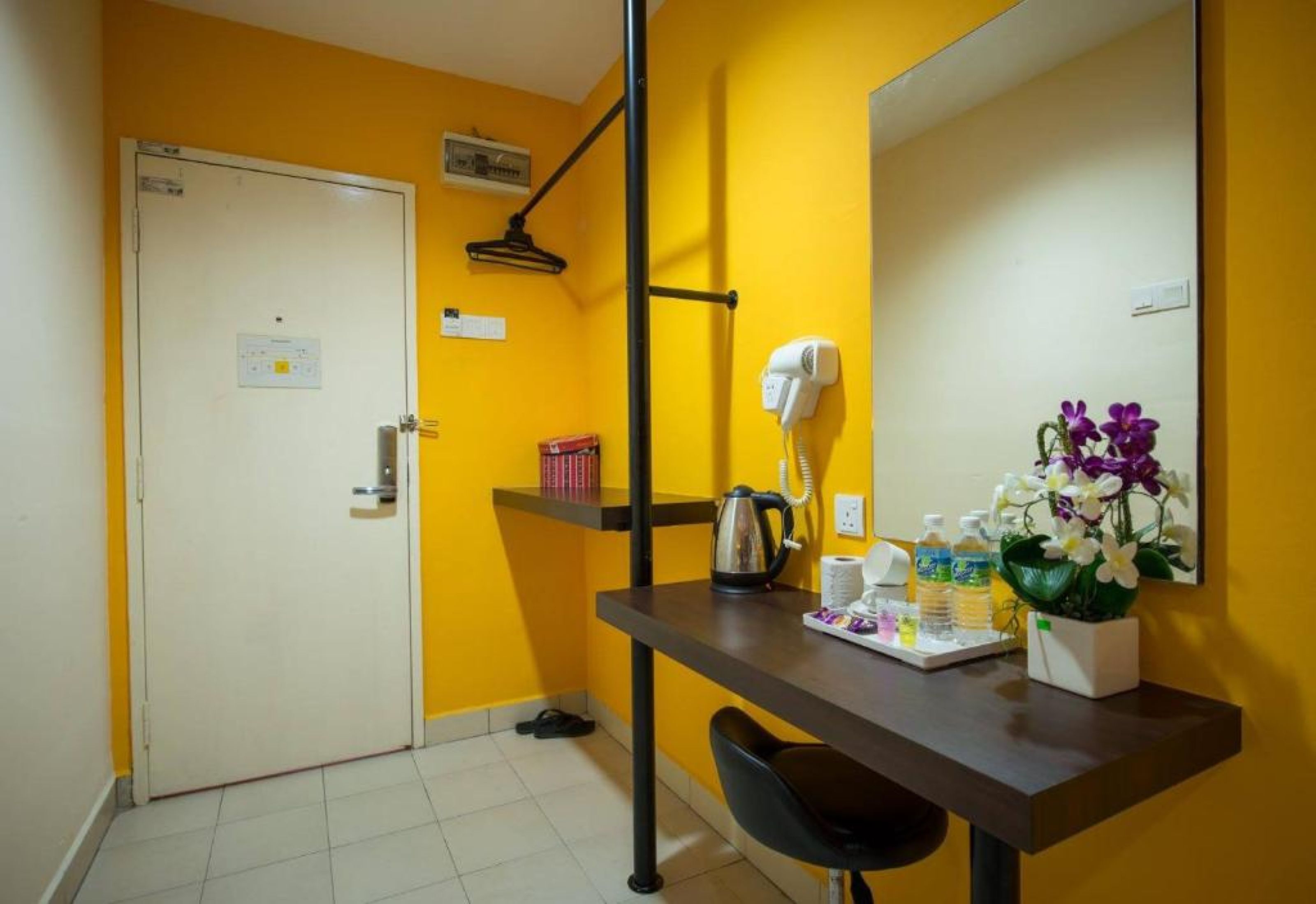 Habitación de hotel con pared amarilla, puerta blanca, escritorio con secador de pelo, hervidor y una planta.