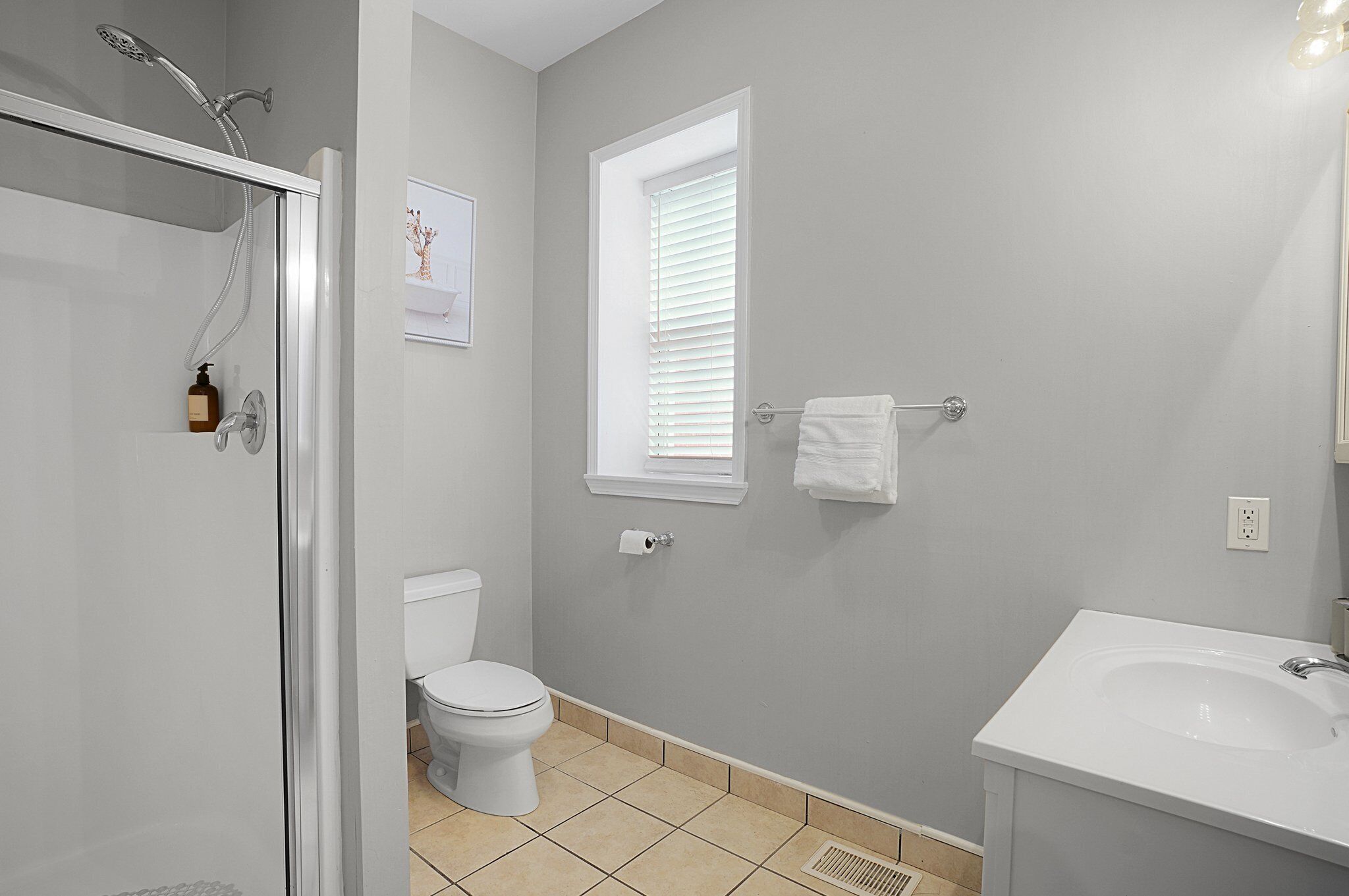 A bathroom with a shower, toilet, sink, and a window with blinds.
