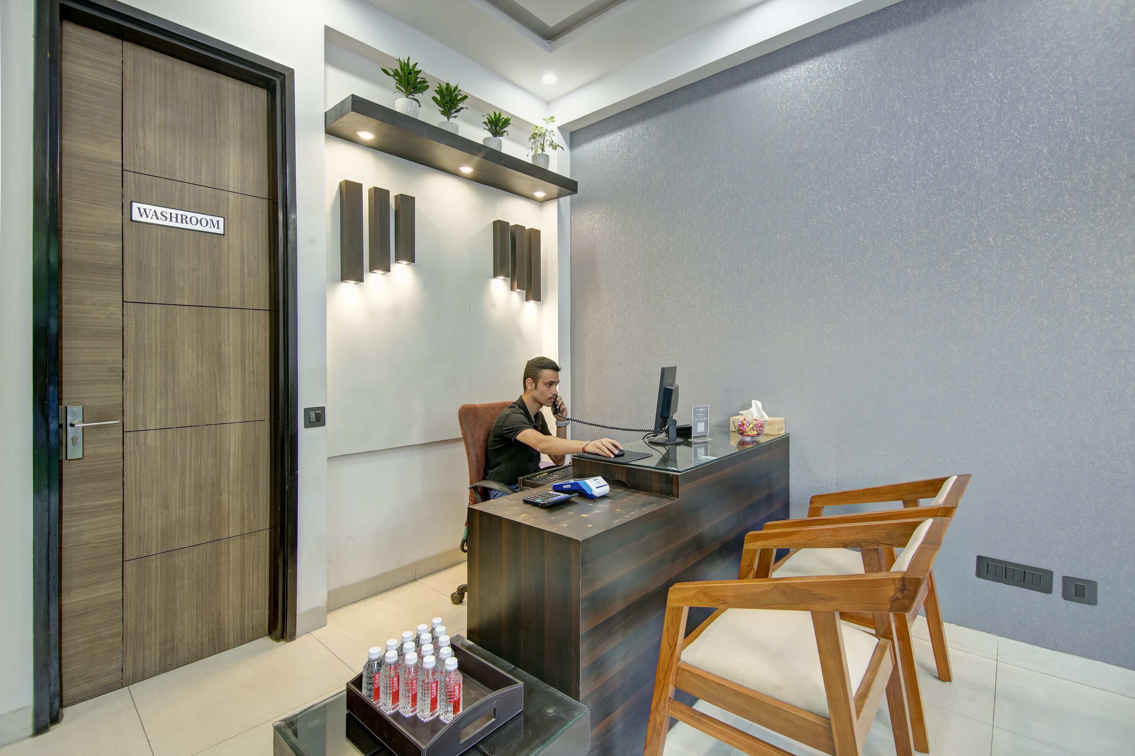 A reception desk with a computer, a phone, and a water dispenser. There are two wooden chairs and a small table with water bottles. A sign on the door reads "WASHROOM".