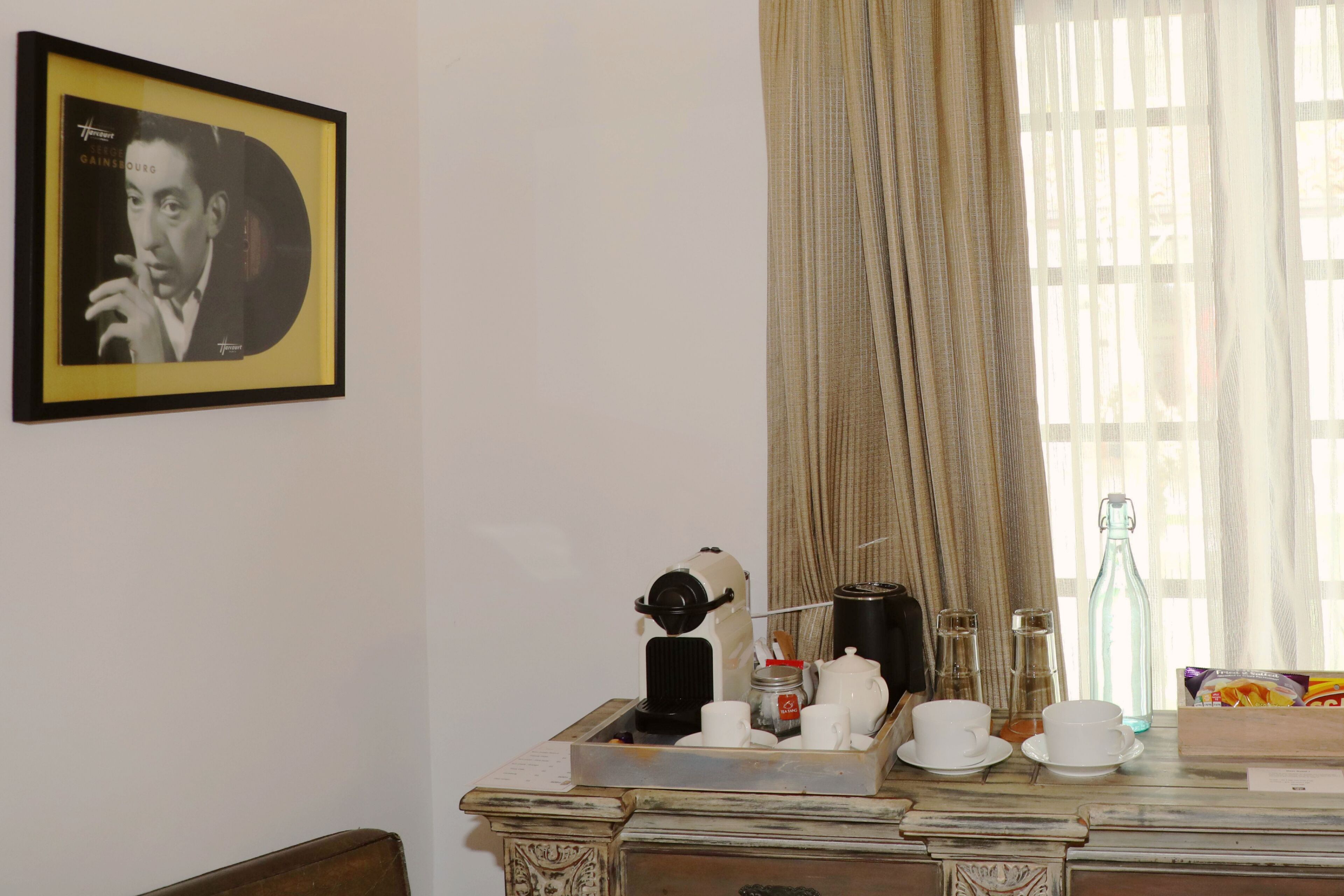 A room with a framed picture on the wall, a window with curtains, a table with cups, a teapot, a coffee maker, and a bottle.