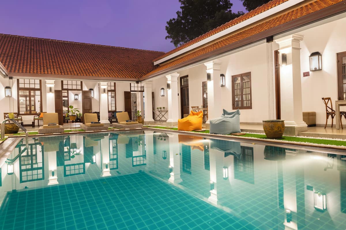 Piscina com deck revestido em azulejo, área de estar coberta com cadeiras e mesas, e um edifício com telhado e paredes brancas.