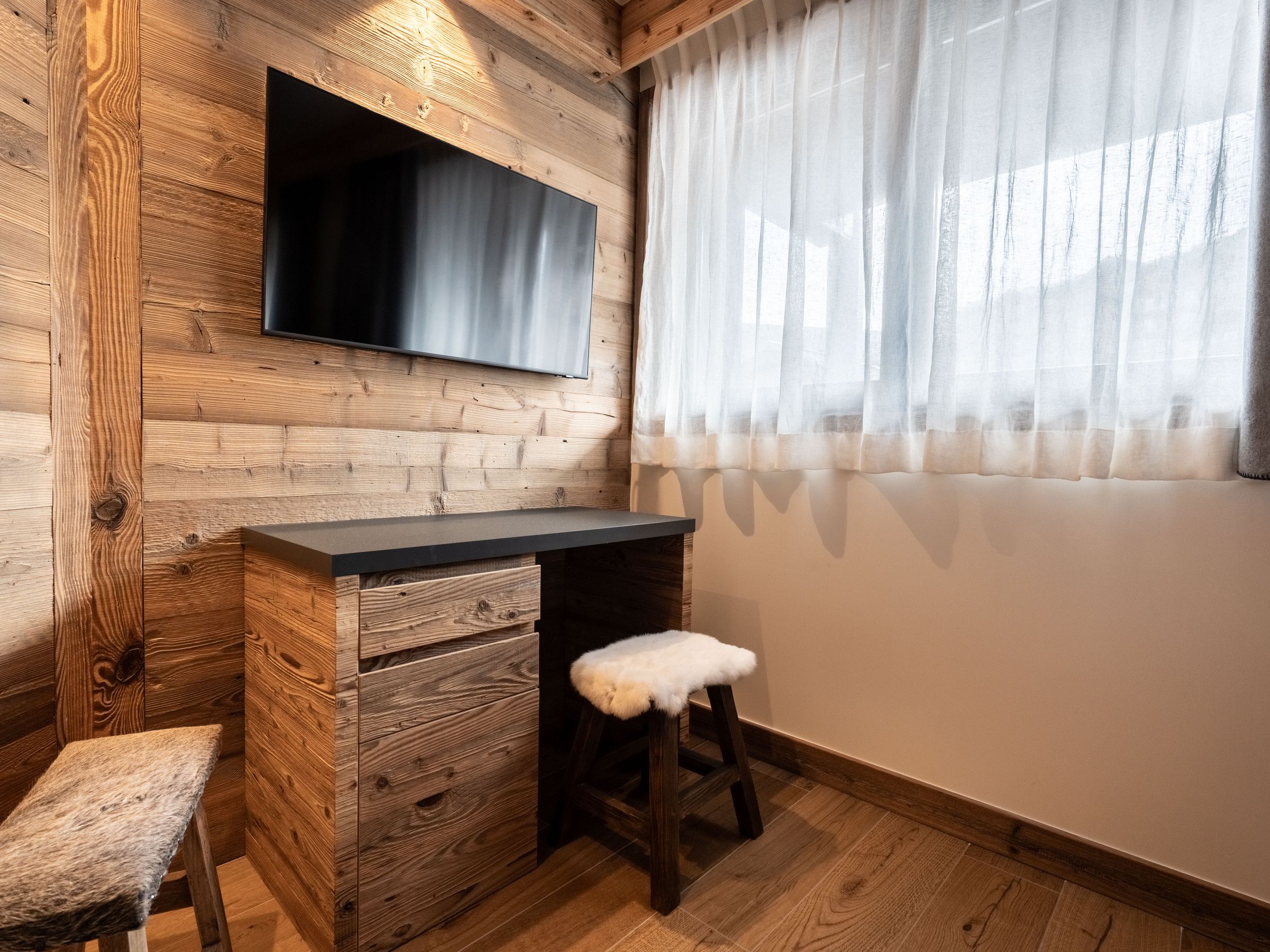 Ein gemütliches Zimmer mit einer Holzwand, einem Flachbildfernseher über einer dunklen Arbeitsfläche, einem kleinen Hocker mit Fellbezug und einem Fenster mit durchscheinenden Vorhängen.