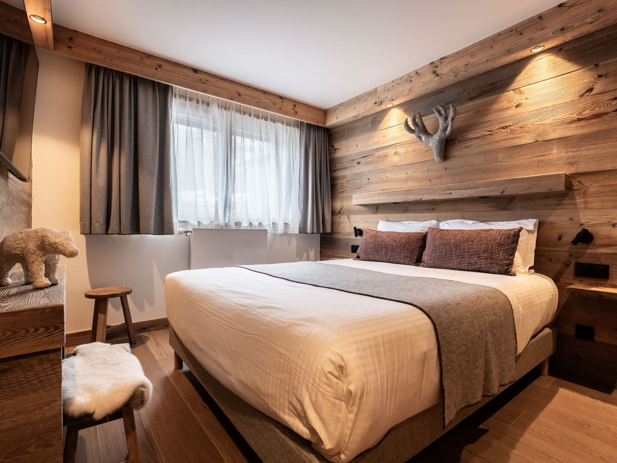 Une chambre à coucher avec une tête de lit en bois, un lit avec une literie blanche, un petit tabouret et une statue d’ours.