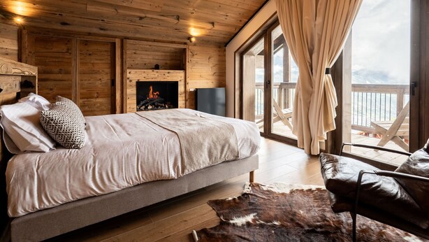 A cozy cabin-style bedroom with a bed, a fireplace, a large window with curtains, and a wooden ceiling.