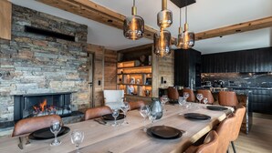 A modern dining room with a long wooden table set for a meal, a stone fireplace, and a kitchen area in the background.