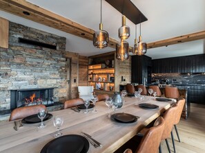 A modern dining room with a long wooden table set for a meal, a stone fireplace, and a kitchen area in the background.
