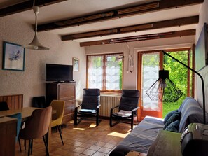 Uma sala de estar acolhedora com televisão, vigas de madeira no teto, área de refeições com cadeiras e grandes janelas com vista para o verde lá fora.