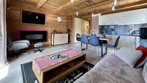 Tunnelmallinen mökin sisustus, jossa on puinen katto, seinälle asennettu televisio, ruokailutila tuoleineen ja pöytineen sekä keittiö kaappeineen ja kodinkoneineen.