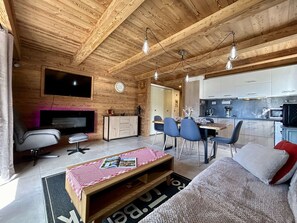 A cozy cabin interior with a wooden ceiling, a TV mounted on the wall, a dining area with chairs and a table, and a kitchen with cabinets and appliances. - Cozy apartment with balcony and parking in Briançon (Briançon)