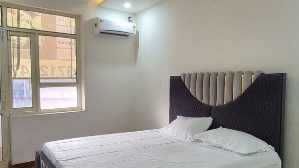 A bedroom with a bed, ceiling fan, air conditioning unit, and a window. - Ambika Hotels (Faridabad)